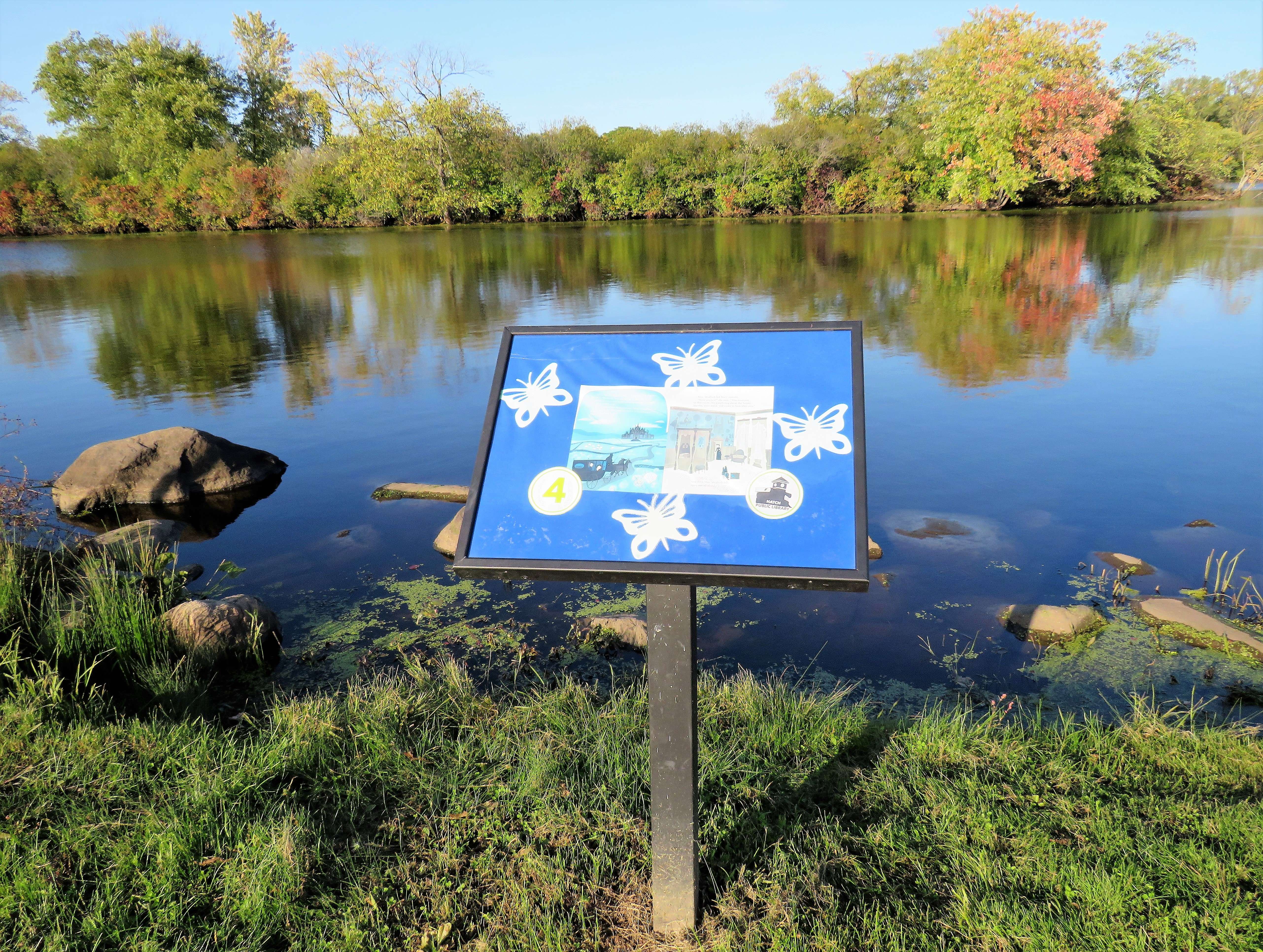 An angled StoryWalk frame on a single post beside a pond